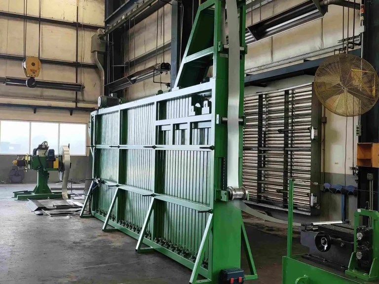 A tall vertical coil accumulator (loop pit or tower accumulator) designed to store a buffer of material, allowing the roll forming machine to run continuously while new coils are being joined or prepared.